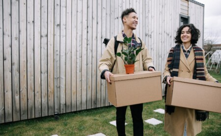 a happy couple carrying cardboard boxes and backpacks entering their new homes' yard