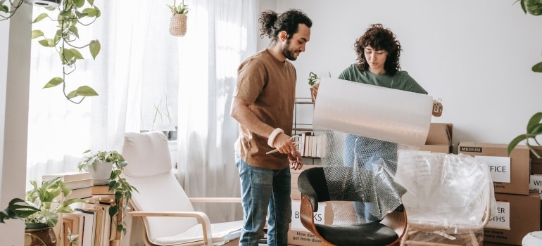 a couple placing bubble wrap around a wooden chair as one of the tips to reduce house moving costs
