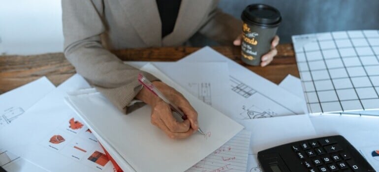 A woman holding a coffee while writing calculations in the notebook