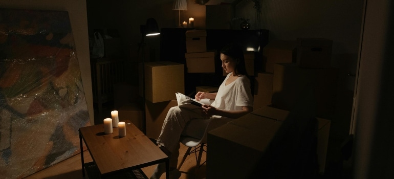Woman reading a book in a dimly lit room surrounded by moving boxes and candlelight.