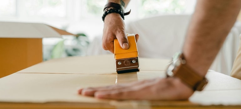 Close-up of a person sealing a cardboard box with tape, showcasing sustainable packing practices in a bright indoor setting.