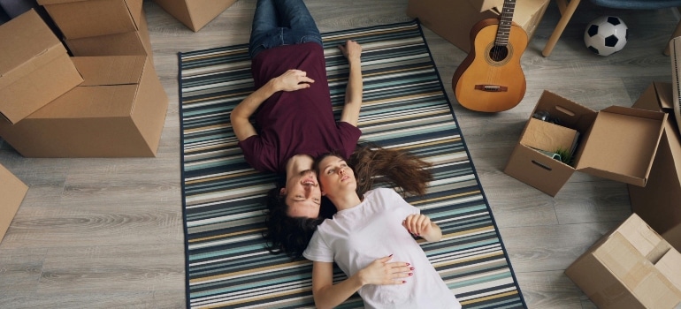 Couple relaxing on a rug surrounded by unpacked boxes and enjoying their first day in the new home.