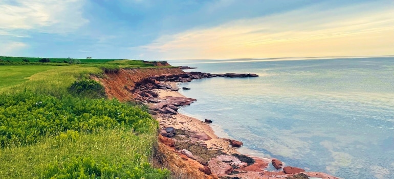 Rocky shoreline on Prince Edward Island with green fields and calm ocean at sunset.