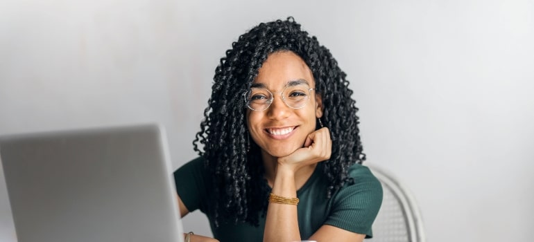 Smiling young woman with curly hair and glasses working on a laptop.